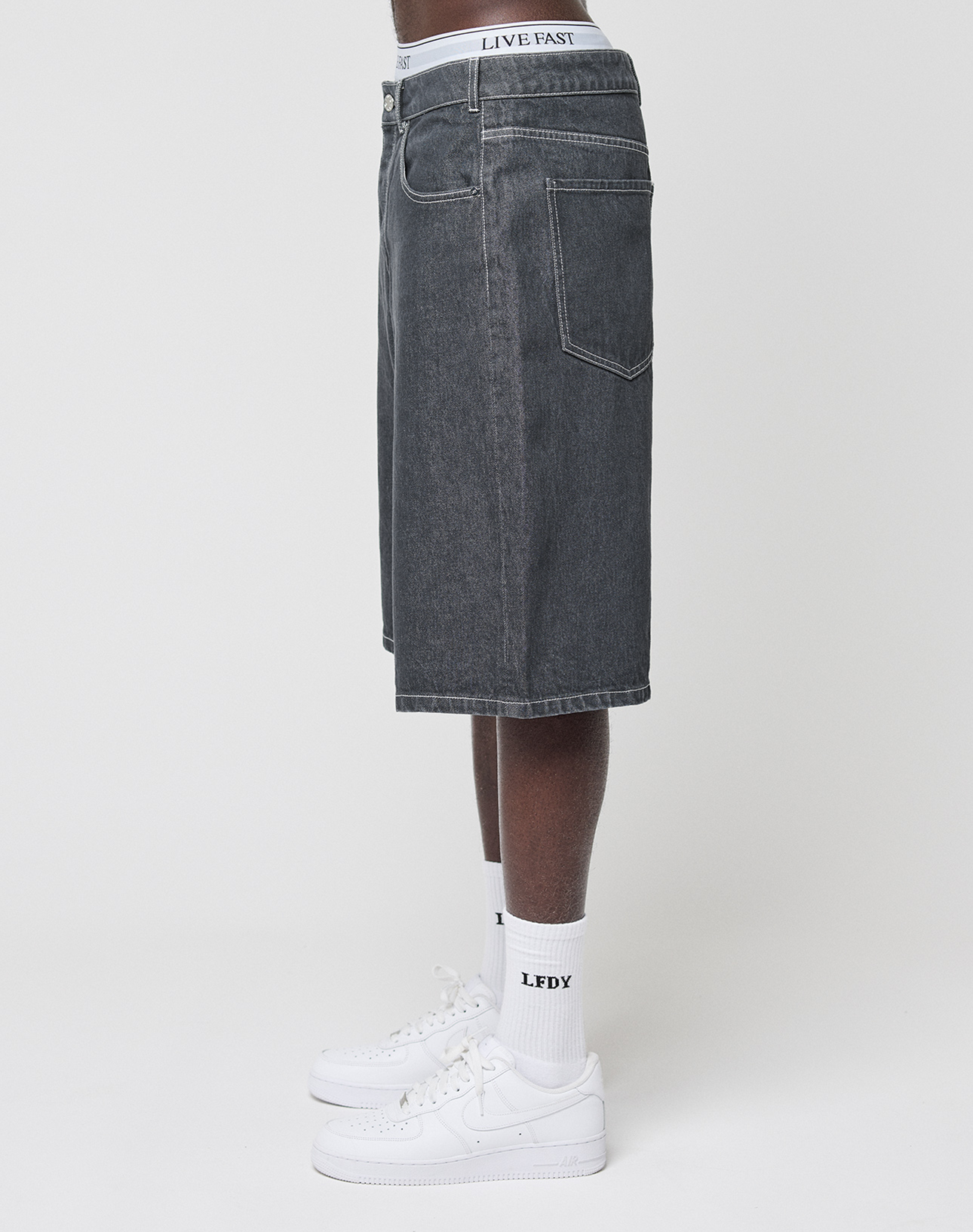 A person stands sideways against a white background wearing baggy fit LFDY Jorts by LIVE FAST DIE YOUNG, featuring a subtle Lederpatch, a white LIVE FAST waistband, LFDY-branded white socks, and white sneakers.