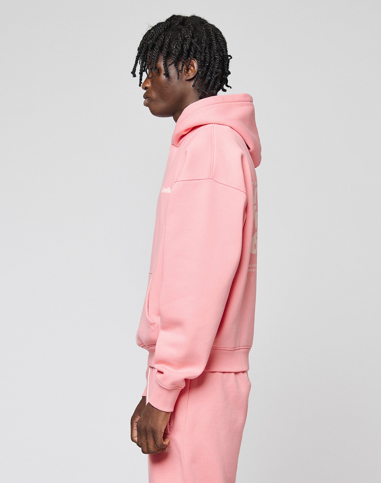 A person with short, twisted hair stands sideways, wearing a pink LIVE FAST DIE YOUNG Basic 360 Hooded sweatshirt and matching sweatpants with topstitching, against a plain light gray background.