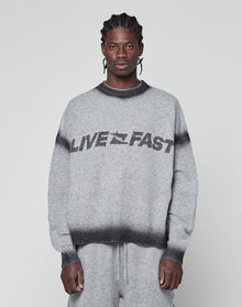 A man with braided hair wears an oversize fit grey Live Fast Boucle Sweater by LIVE FAST DIE YOUNG, featuring a dark faded spray effect stripe and bold LIVE FAST lettering, paired with matching pants against a plain light background. | model