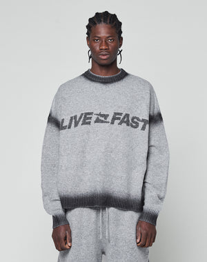 A man with braided hair wears an oversize fit grey Live Fast Boucle Sweater by LIVE FAST DIE YOUNG, featuring a dark faded spray effect stripe and bold LIVE FAST lettering, paired with matching pants against a plain light background.