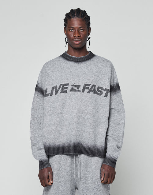 A man with braided hair wears an oversize fit grey Live Fast Boucle Sweater by LIVE FAST DIE YOUNG, featuring a dark faded spray effect stripe and bold LIVE FAST lettering, paired with matching pants against a plain light background.