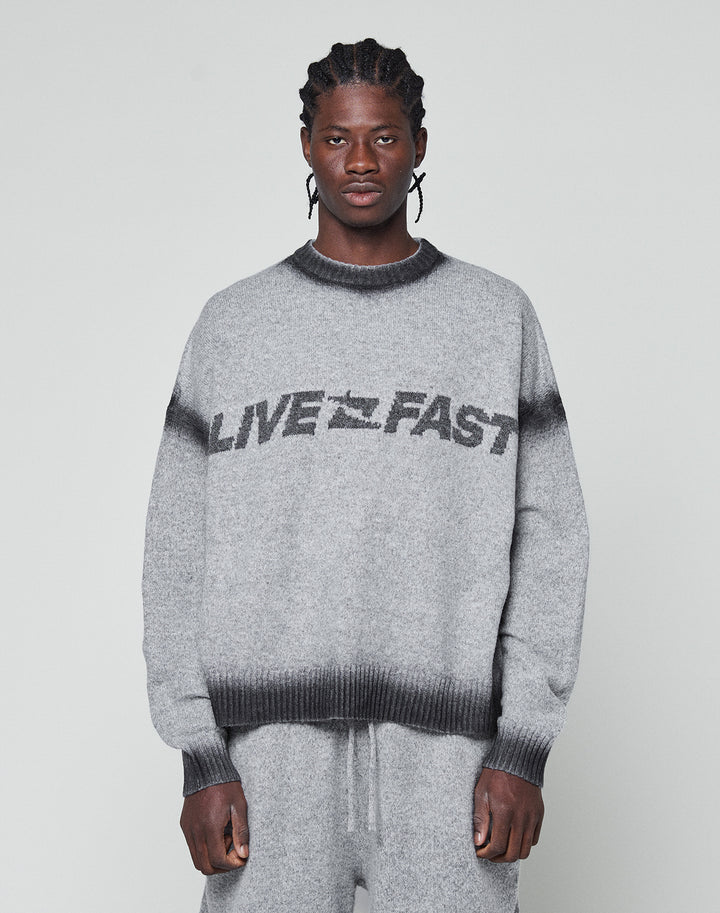 A man with braided hair wears an oversize fit grey Live Fast Boucle Sweater by LIVE FAST DIE YOUNG, featuring a dark faded spray effect stripe and bold LIVE FAST lettering, paired with matching pants against a plain light background. | model
