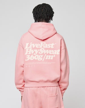 A person with short, twisted hair wears a pink LIVE FAST DIE YOUNG Basic 360 Hooded and matching pants. The hoodie has bold front and back print, sturdy topstitching, and white text. The background is plain.