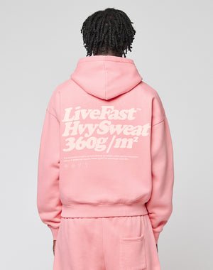 Person with short, twisted hair facing away, wearing an Oversize Fit pink Basic 360 Hooded and pants by LIVE FAST DIE YOUNG. The hoodie has bold white "LiveFast HvySweat 360g/m²" text and visible topstitching on the back. Background is plain light gray.