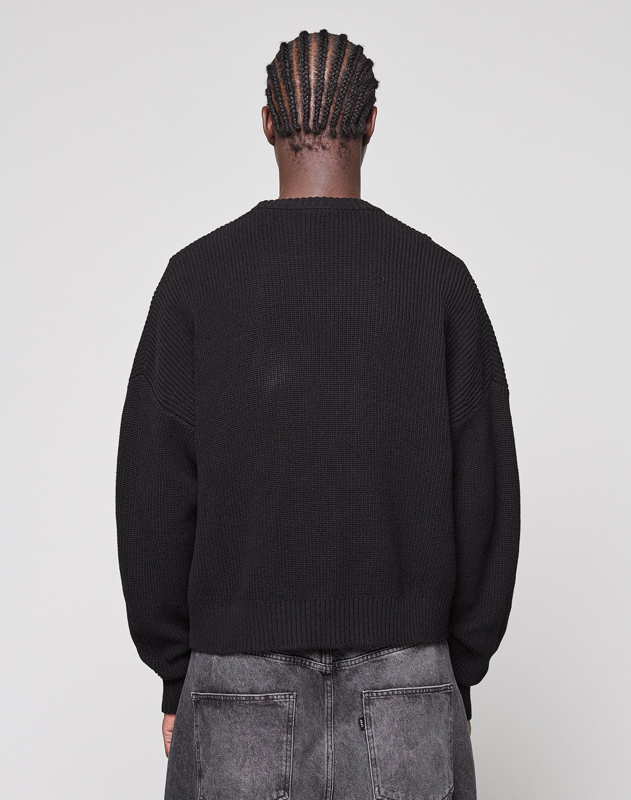 A person with cornrowed hair faces away from the camera, wearing a loose-fit LFDY Knit Sweater by LIVE FAST DIE YOUNG and faded black jeans against a plain light gray background.