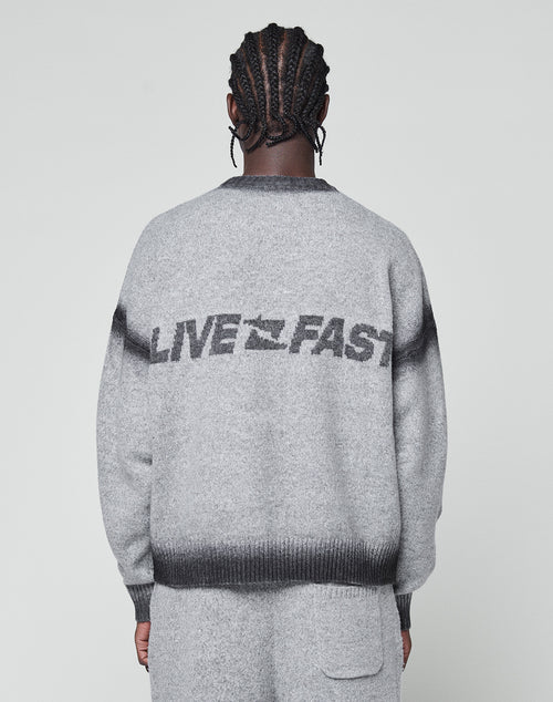 A person with braided hair, seen from behind, wears the LIVE FAST DIE YOUNG Live Fast Boucle Sweater in light gray. "LIVE FAST" and a bird graphic are on the back with a faded spray effect and dark gray stripes at shoulders and hem.