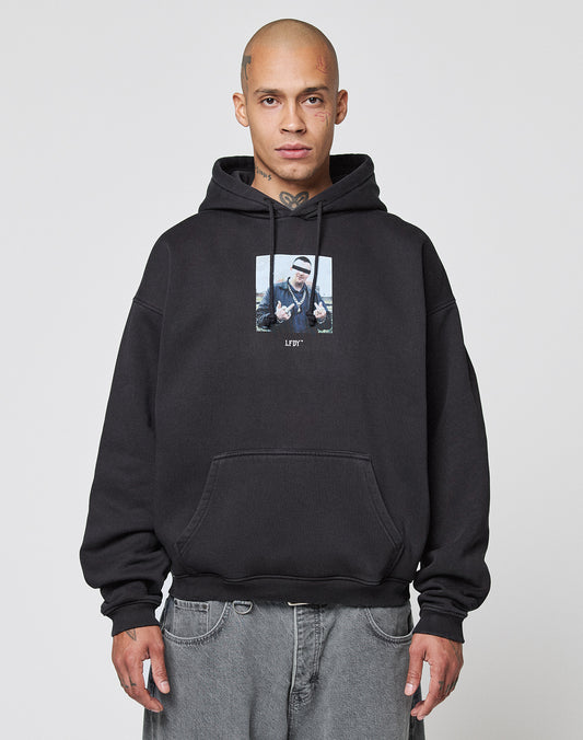 A person with a shaved head and tattoos wears the oversize black LFDY Gambling Hooded by LIVE FAST DIE YOUNG, featuring a bold front print. Standing forward with relaxed hands, they pair it with faded gray jeans.
