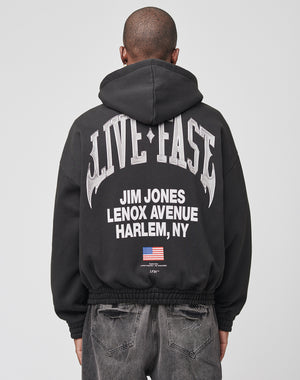 A person faces away from the camera wearing a black, oversized LIVE FAST DIE YOUNG Live Fast Lenox Hooded Zip, made of Baumwolle, with bold front and back prints including JIM JONES LENOX AVENUE HARLEM, NY and a small American flag on the back.