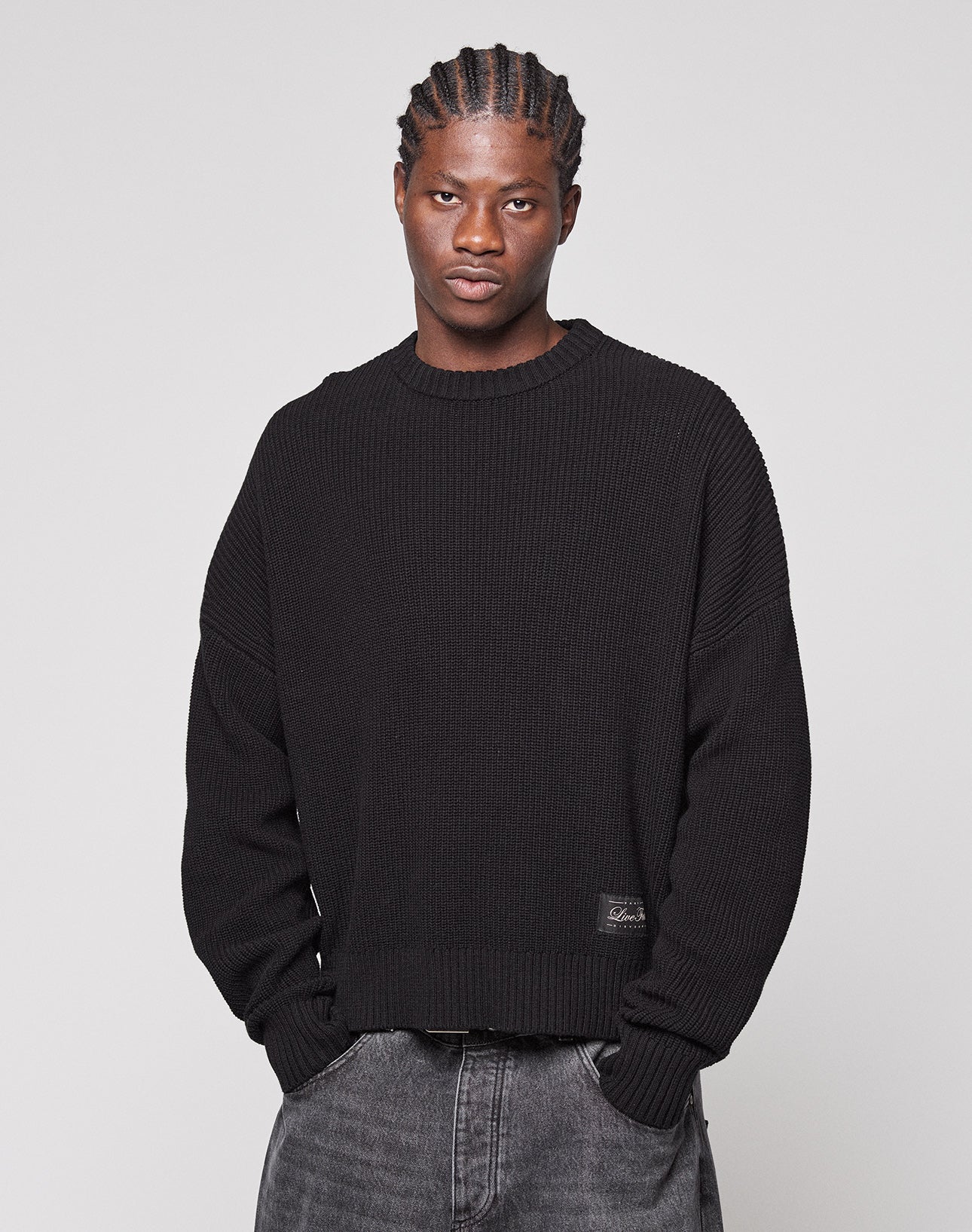 A person with braided hair wears the LFDY Knit Sweater by LIVE FAST DIE YOUNG—an oversized, loose black 100% Baumwolle (cotton) knit with dropped shoulders—paired with faded gray jeans, standing against a plain light gray background.
