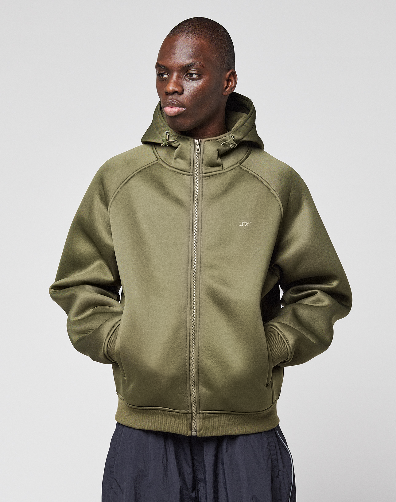 Someone wears an olive green LFDY Scuba Hooded Zip by LIVE FAST DIE YOUNG, hood up and hands in zip pockets, standing against a plain light gray background.