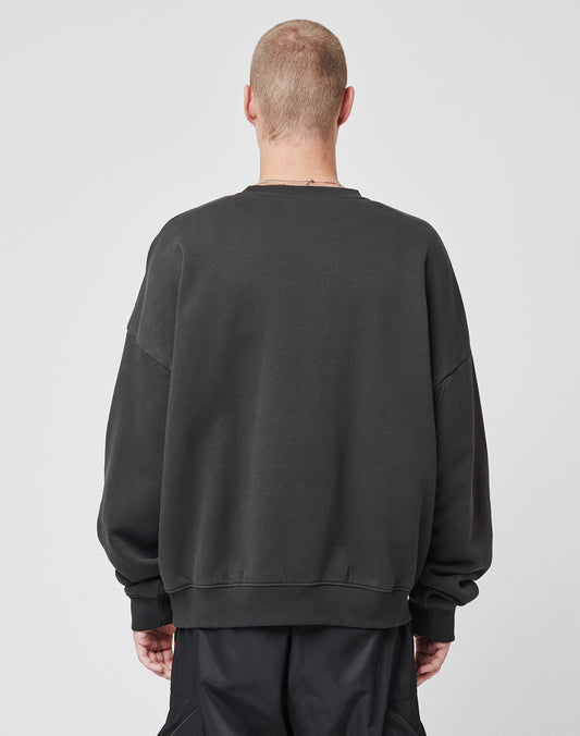 A person with short hair stands with their back to the camera, wearing the LIVE FAST DIE YOUNG Live – Fast Sweater, an oversized dark soft Baumwolle sweatshirt, paired with black pants against a plain light background.