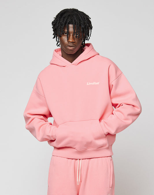A person with curly hair and a neutral expression wears the LIVE FAST DIE YOUNG Basic 360 Hooded in an oversize pink fit with matching sweatpants. The hoodie features white "Livefast" chest text, visible topstitching, and soft Baumwolle fabric.