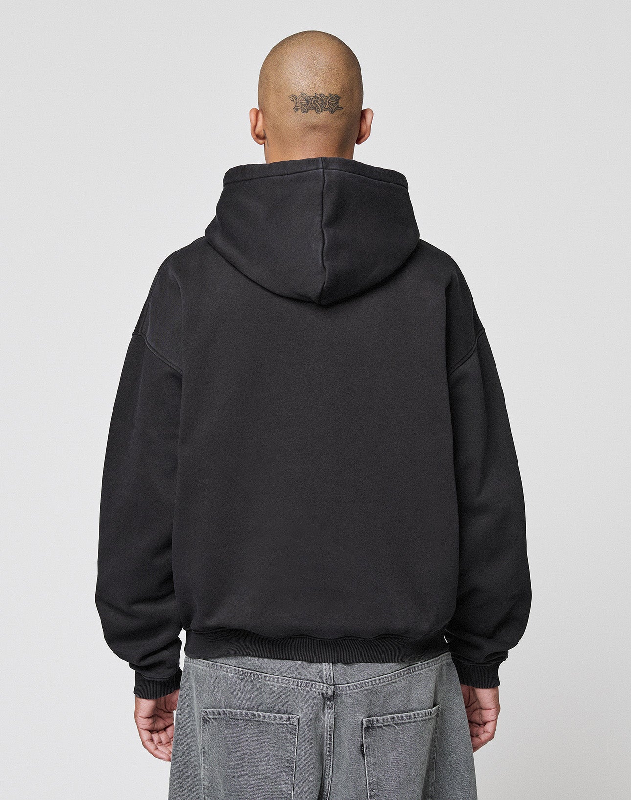 A person with a shaved head and a head tattoo is seen from behind, wearing the black LIVE FAST DIE YOUNG LFDY Gambling Hooded (360 GSM, oversize fit) and gray jeans against a plain light background.