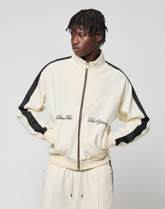 Someone stands against a plain background wearing the cream-colored LFDY Striped Track Jacket by LIVE FAST DIE YOUNG, featuring raglan sleeves, black stripes on the arms, and "Live Fast and Die Young" embroidered on the front.