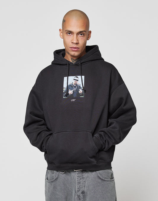 A person with a shaved head faces the camera against a plain light background, wearing gray jeans and a black LFDY Gambling Hooded sweatshirt by LIVE FAST DIE YOUNG, both with fine topstitching and hands in their pockets.