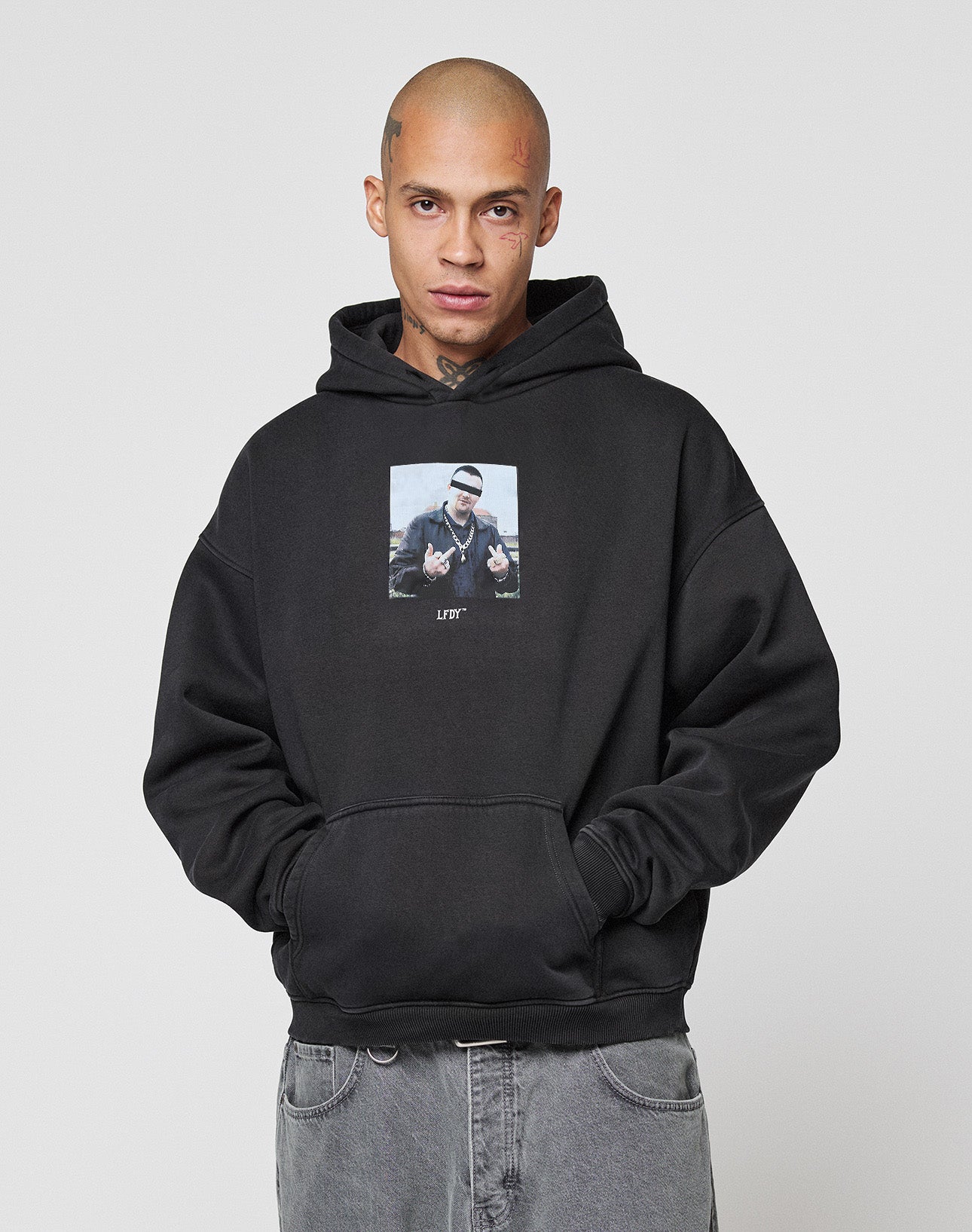 A person with a shaved head, tattoos, and neutral expression wears the LIVE FAST DIE YOUNG LFDY Gambling Hooded—a black oversize 360 GSM hoodie with a bold front photo print—standing against a plain light gray background with hands in the pocket.