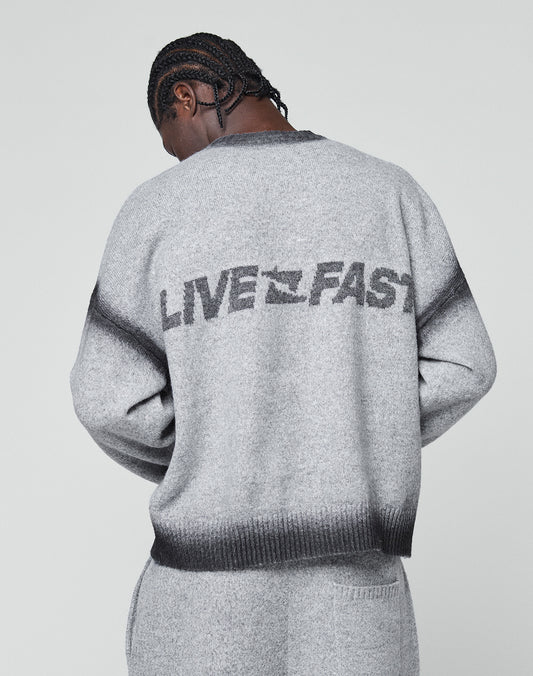 A person with braided hair faces away, wearing the LIVE FAST DIE YOUNG Live Fast Boucle Sweater—an oversize gray piece featuring bold “LIVE FAST” lettering on the back. The background is plain and light-colored.