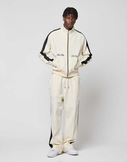 A man stands against a white background wearing the LIVE FAST DIE YOUNG LFDY Striped Track Jacket with black sleeve stripes and matching pants. He has short dreadlocks and white sneakers.