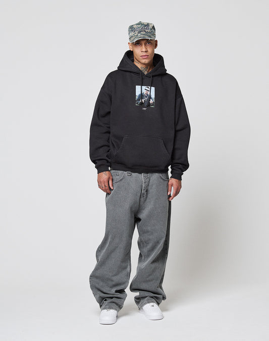 A man wearing a camo cap, LIVE FAST DIE YOUNG LFDY Gambling Hooded with frontprint, baggy gray topstitched jeans, and white sneakers stands against a plain white background.
