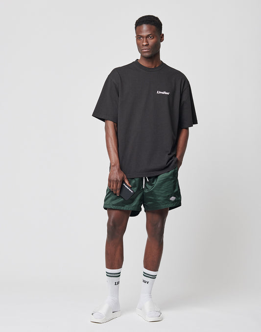 A man stands against a plain background wearing a black oversized t-shirt, LIVE FAST DIE YOUNG Basic Satin Shorts in green camo with Elastikbund, white socks with black stripes, white slide sandals, and holds a phone while looking to the side.