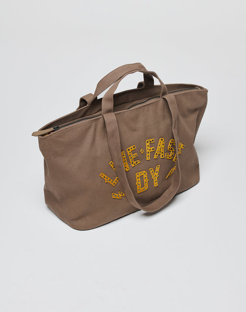 The LFDY Tote Bag by LIVE FAST DIE YOUNG is a brown canvas bag with two short handles, a zipper, a secure laptop compartment, and “THE FAST BOY” in yellow cheetah-print letters on the side, shown on a plain white background.