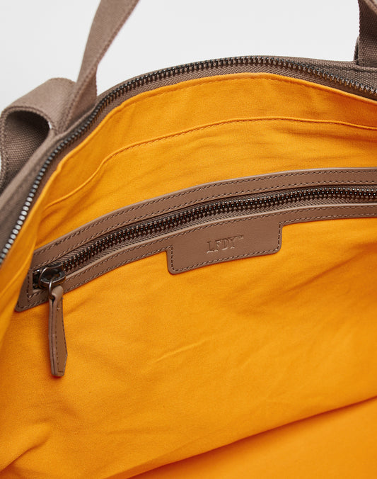 A close-up of the LFDY Tote Bag by LIVE FAST DIE YOUNG shows its orange fabric lining, brown leather-trimmed zippered pocket with LLOY embossing, and a spacious laptop compartment, highlighting quality craftsmanship.