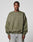 A man in the LFDY Wavy Sweater by LIVE FAST DIE YOUNG, an oversized olive green cotton-poly blend with subtle curved seams and light chest text, pairs it with wide-leg light jeans while standing against a plain gray background.