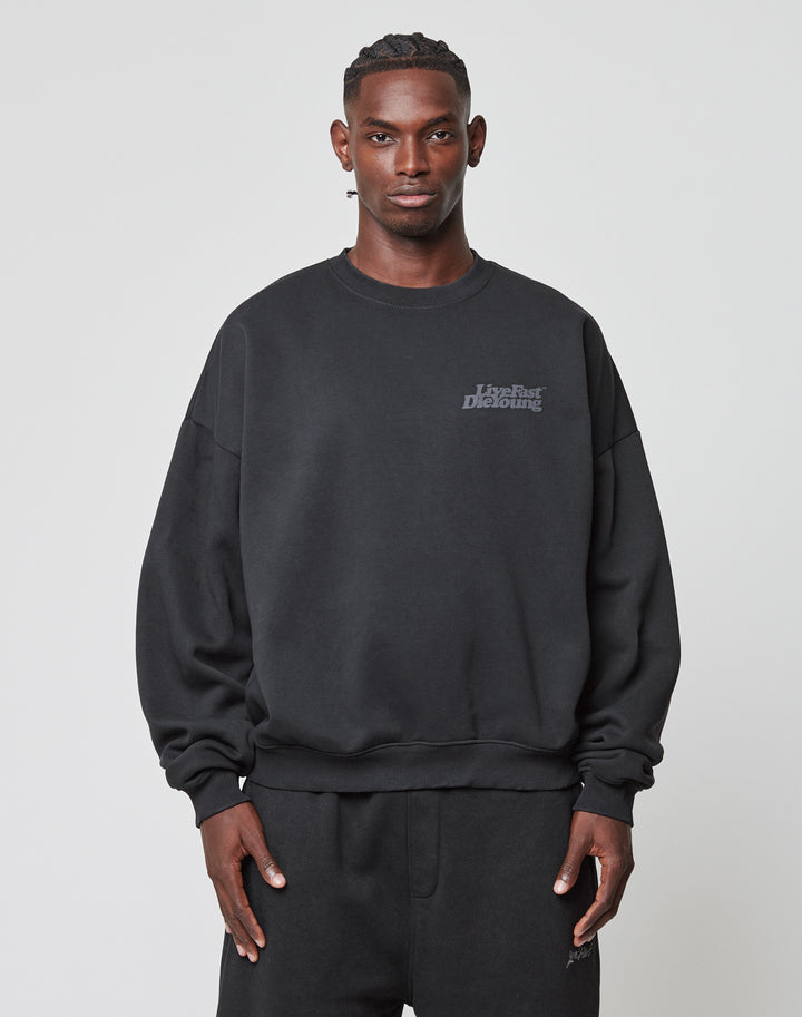 A person faces forward in a black Basic Acid Sweater by LIVE FAST DIE YOUNG, paired with matching black sweatpants, both crafted from a soft cotton-polyester blend, set against a plain light background.