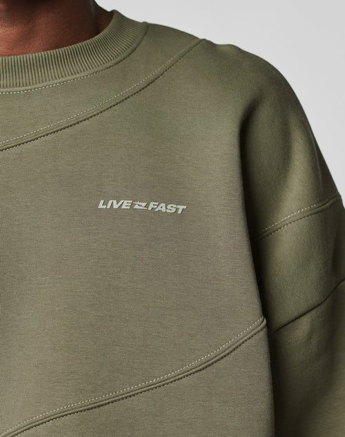 Close-up of a person wearing the LFDY Wavy Sweater by LIVE FAST DIE YOUNG, an olive green oversize fit with LIVE FAST in white on the chest. Made from a soft Baumwolle blend, it has visible stitching and a ribbed neckline.
