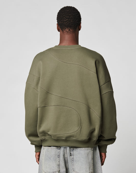 A person with short, textured hair is seen from behind in an LFDY Wavy Sweater by LIVE FAST DIE YOUNG—an oversize olive green top in soft cotton-polyester—paired with light-wash denim jeans against a plain light gray background.
