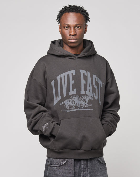 A young man with short dreadlocks wears the Live Fast Stallion Hooded by LIVE FAST DIE YOUNG, an oversize-fit hoodie featuring a tiger front print and LIVE FAST text. He stands with hands in pockets against a plain, light background.
