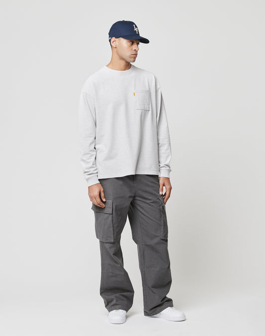 A man in a navy baseball cap, light gray long-sleeve shirt with chest pocket, LIVE FAST DIE YOUNG Wide Cargo dark gray pants with cargo pockets, and white sneakers stands against a plain white background.