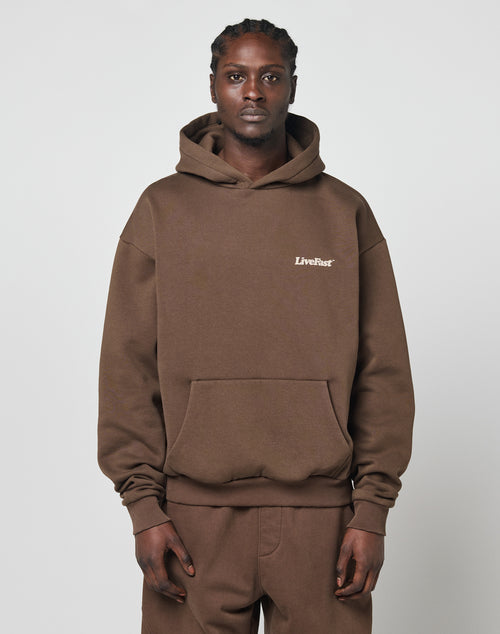 A man stands against a plain light background, wearing a brown LIVE FAST DIE YOUNG Basic 360 Hooded sweatshirt and matching pants. The hoodie features LiveFast printed in small white letters on the chest.
