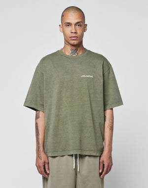 A tattooed person stands against a plain background, wearing the LIVE FAST DIE YOUNG Basic +49 Chest Tee—an olive-green, 100% Baumwolle oversize t-shirt with a #QLLiveFast logo on the left chest—and light-colored mesh shorts.