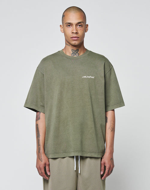 A tattooed person stands against a plain background, wearing the LIVE FAST DIE YOUNG Basic +49 Chest Tee—an olive-green, 100% Baumwolle oversize t-shirt with a #QLLiveFast logo on the left chest—and light-colored mesh shorts.