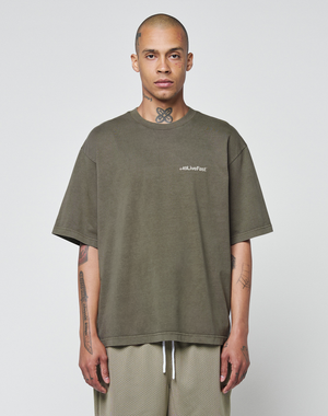 A person with a shaved head and tattoos stands facing forward, wearing the LIVE FAST DIE YOUNG Basic +49 Chest Tee in olive green with logo print and light mesh shorts, set against a plain light background.