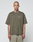 A person with a shaved head and tattoos stands facing forward, wearing the LIVE FAST DIE YOUNG Basic +49 Chest Tee in olive green with logo print and light mesh shorts, set against a plain light background.