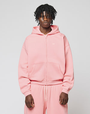 A person with short, curly hair wears the Basic Hooded Zip in light pink by LIVE FAST DIE YOUNG, paired with matching sweatpants featuring subtle branding. They face the camera against a plain light gray background with a neutral expression.
