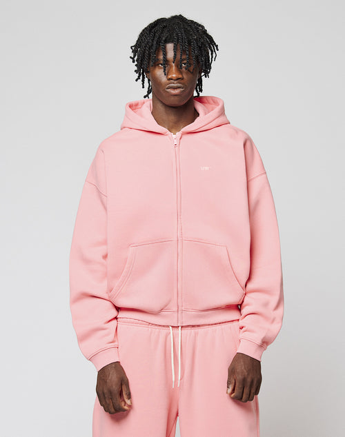 A person with short, curly hair wears the Basic Hooded Zip in light pink by LIVE FAST DIE YOUNG, paired with matching sweatpants featuring subtle branding. They face the camera against a plain light gray background with a neutral expression.
