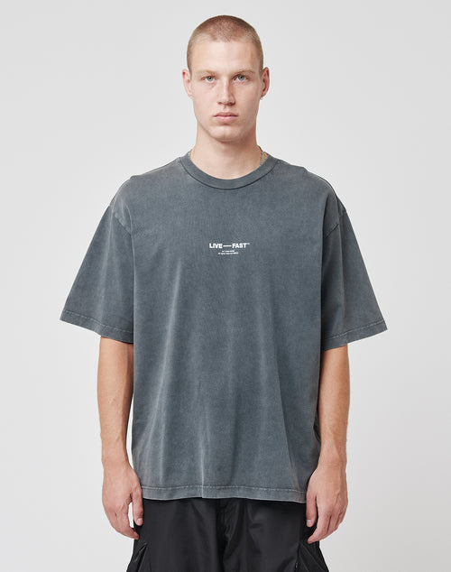A person with closely cropped hair wears the faded black Its Vulgar Tee by LIVE FAST DIE YOUNG, made of 100% Baumwolle with LIVE — FAST in white and subtle front and back prints, standing against a plain white background.