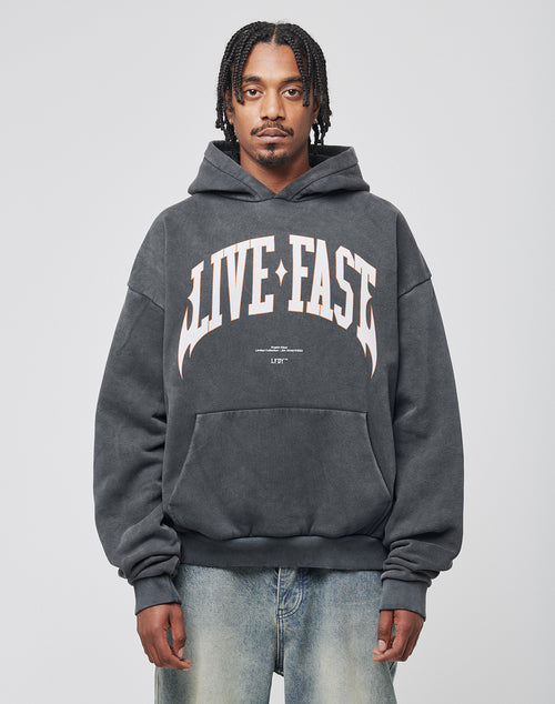 A man with braided hair models the LIVE FAST DIE YOUNG Live Fast Axe Hooded, a soft Baumwolle oversize-fit dark gray hoodie with bold white LIVE FAST lettering, paired with light blue jeans against a plain light background.