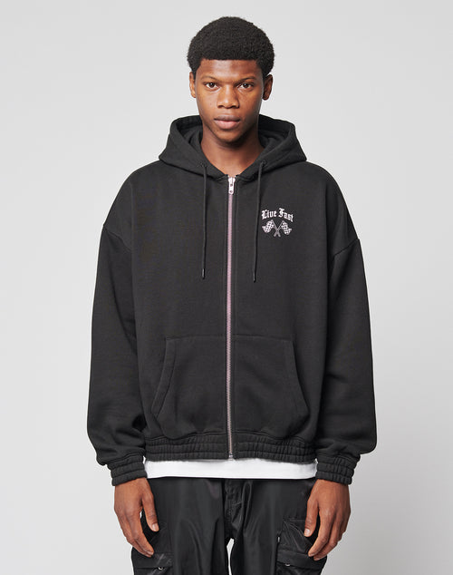 A young man wears the LIVE FAST DIE YOUNG LFDY Haters Hooded Zip—an oversize black zip-up hoodie with a white graphic and text on the left chest—while facing forward against a plain light gray background.