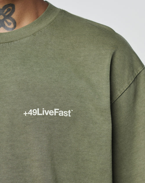 Someone wearing the olive green LIVE FAST DIE YOUNG Basic +49 Chest Tee, featuring a white +49LiveFast logo on the chest. The 100% Baumwolle tee has an oversize fit, with part of a neck tattoo visible at the top of the image.