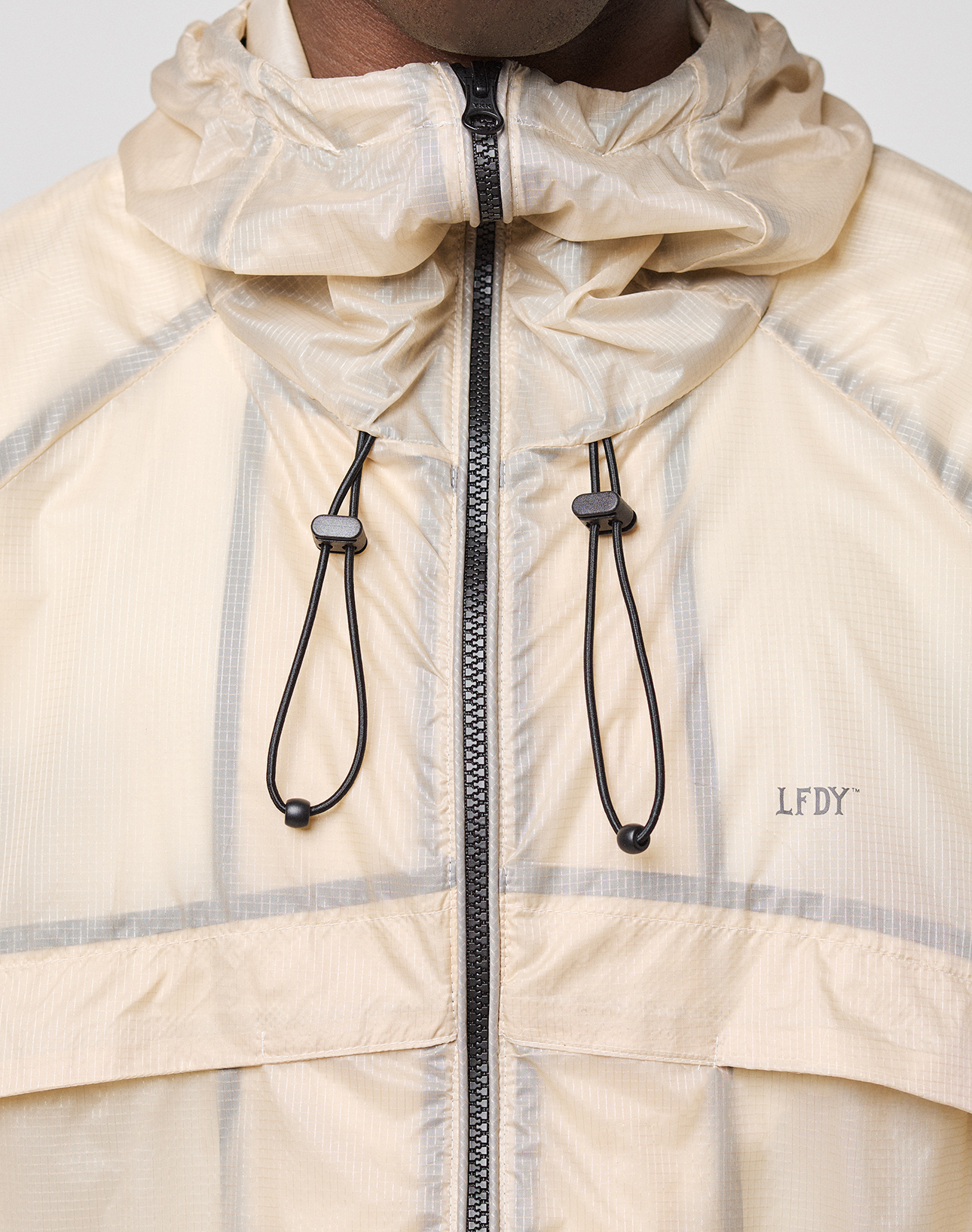 Close-up of a person wearing the beige Live Fast Nylon Jacket by LIVE FAST DIE YOUNG, featuring black drawstrings, a zipper, LFDY logo on the left chest, and a transparent outer layer. Only the lower face and neck are visible.