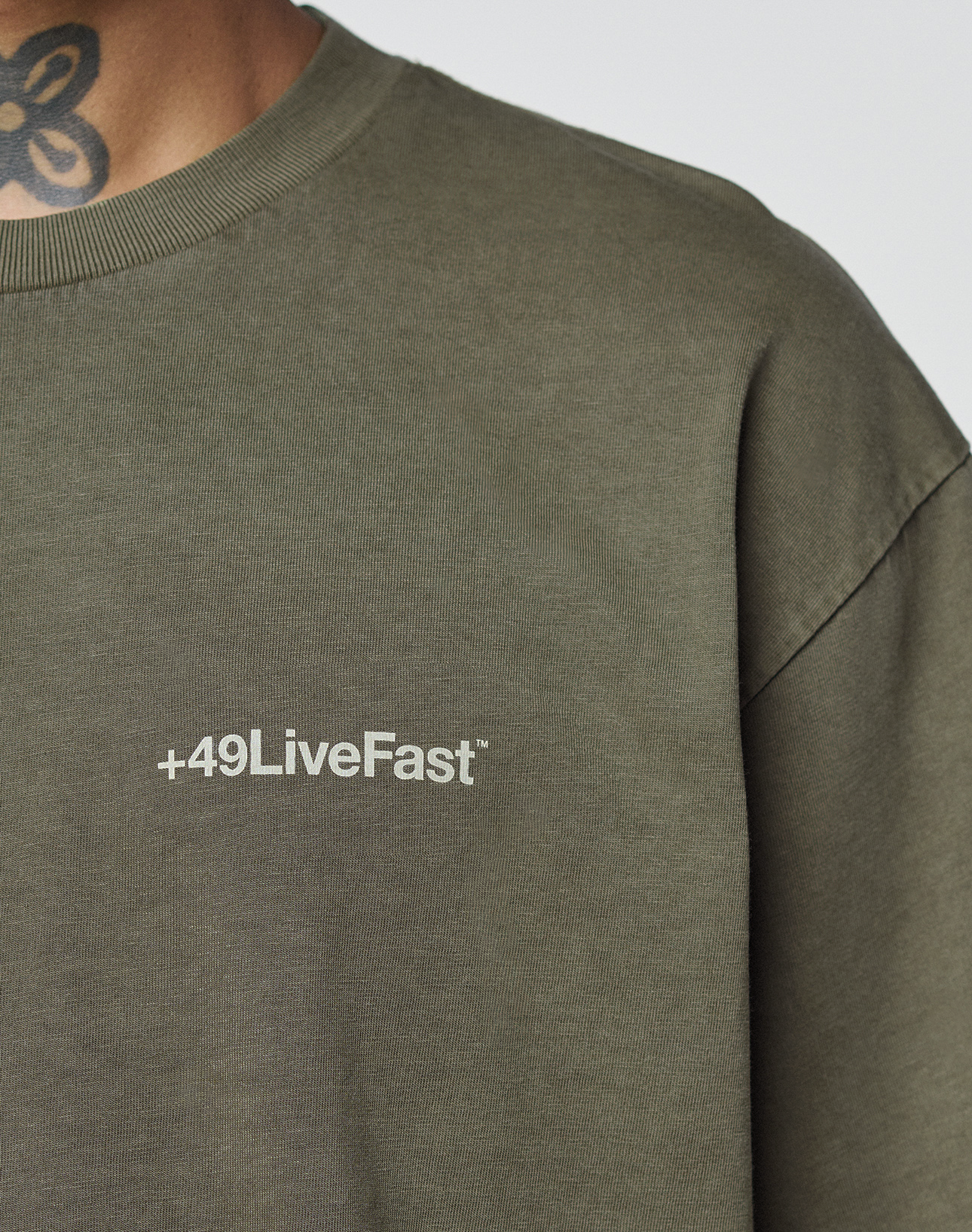 Close-up of a person in an olive green LIVE FAST DIE YOUNG Basic +49 Chest Tee with a white logo on the chest. Made from 100% cotton in an oversize fit, the shirt shows part of a floral neck tattoo above the collar.