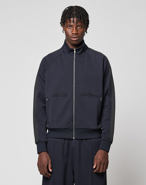 A man with short braided hair wears the LIVE FAST DIE YOUNG LFDY Striped Track Jacket with matching pants, standing against a plain light gray background and looking directly at the camera with a neutral expression.