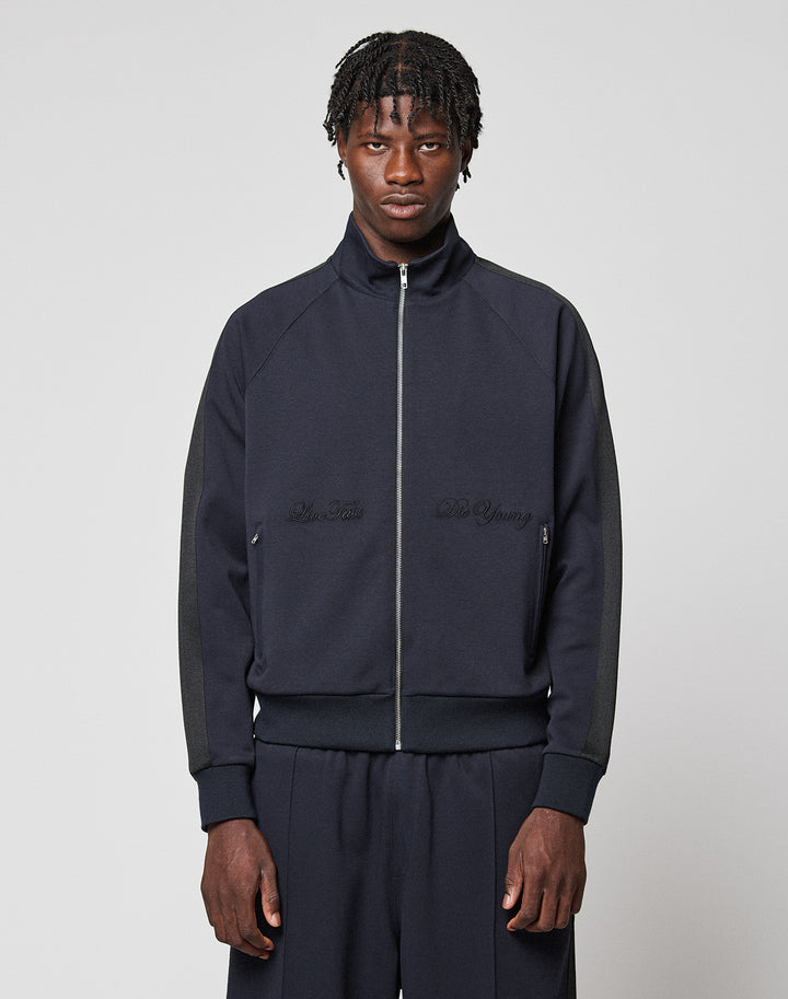 A man with short braided hair wears the LIVE FAST DIE YOUNG LFDY Striped Track Jacket with matching pants, standing against a plain light gray background and looking directly at the camera with a neutral expression. | model