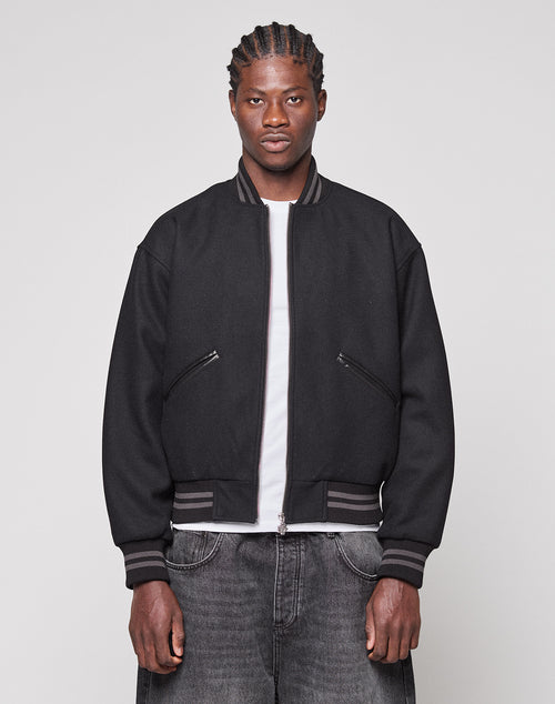 A person with braided hair wears the LIVE FAST DIE YOUNG Live Fast College Jacket, featuring a metal zipper, over a white T-shirt and dark jeans, standing against a plain light gray background and looking directly at the camera.