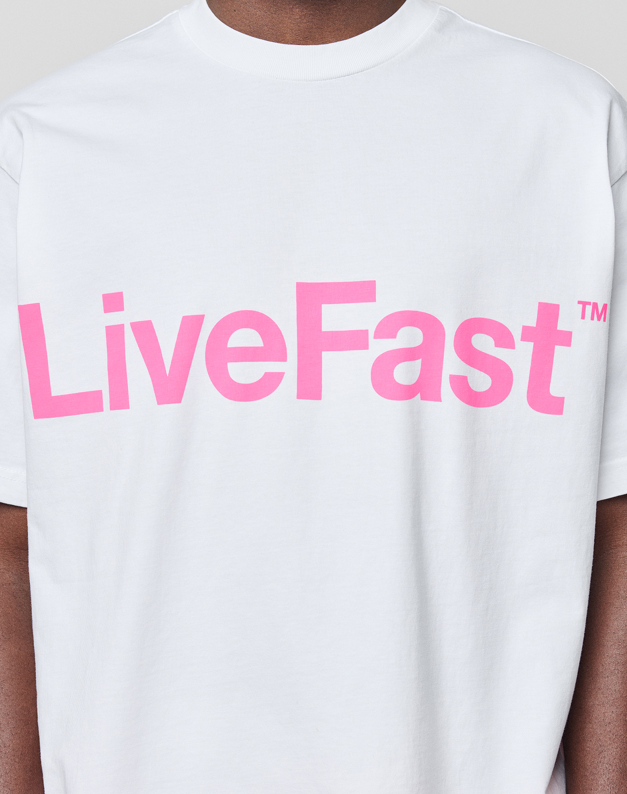 Someone wears the Basic OS Livefast Tee by LIVE FAST DIE YOUNG—an oversized white t-shirt made from 100% Baumwolle, featuring a bold pink LiveFast™ print across the chest.