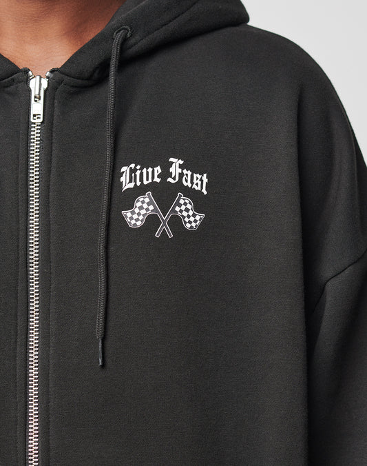 A person wears the LIVE FAST DIE YOUNG LFDY Haters Hooded Zip, an oversized black zip-up hoodie with Live Fast and two crossed checkered flags in white on the left chest. Only the upper body is shown.